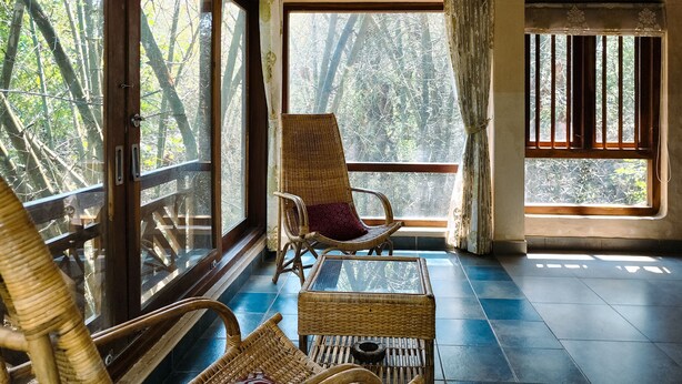 Ein Zimmer mit großen Fenstern, Korbmöbeln und Blick auf Bäume.