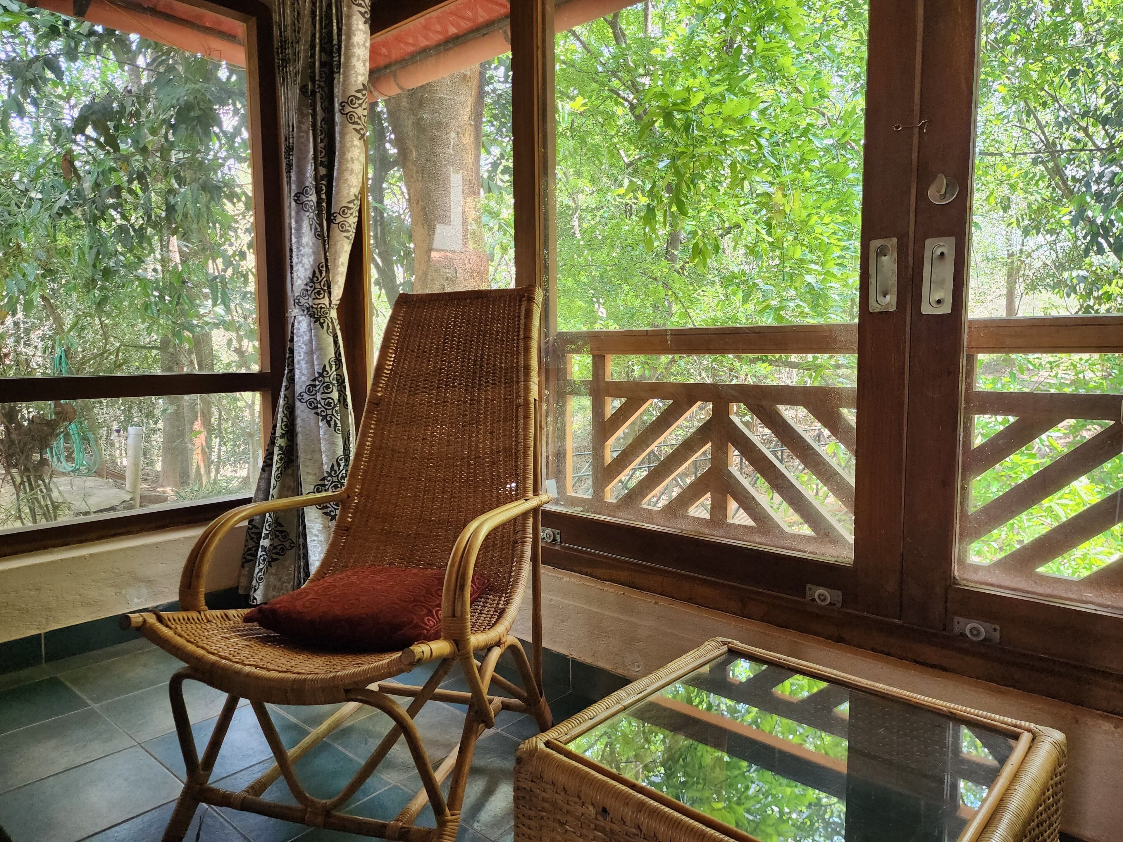 Une chaise en osier avec un coussin rouge, placée sur un balcon où se trouve une table en verre, le tout offrant une vue sur une forêt verdoyante.