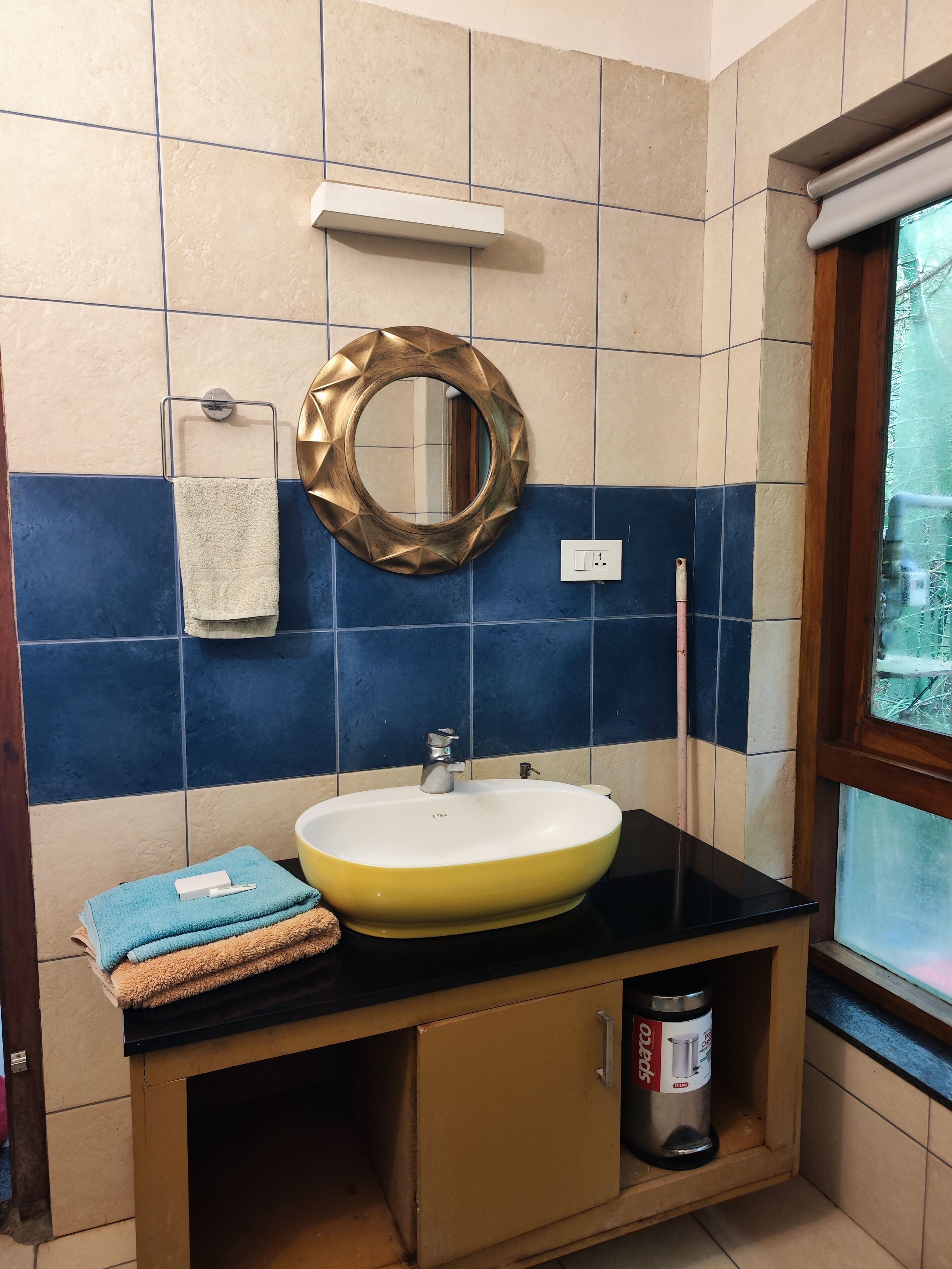 A bathroom with a yellow sink, a mirror with a gold frame, a towel rack, and a window with a view of trees.