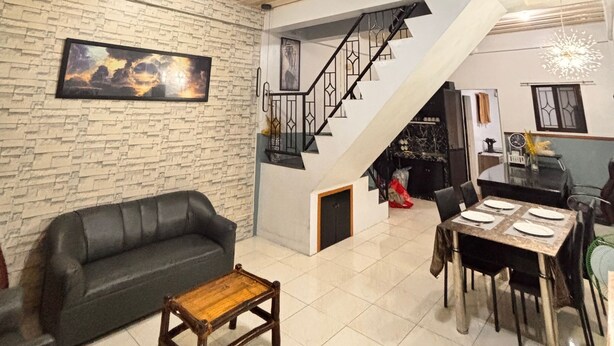 A spacious living area with a black leather sofa, a wooden coffee table, a dining table set for four, and a staircase leading to an upper level.
