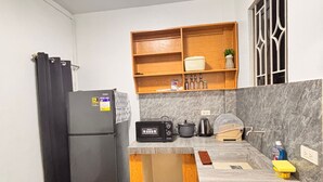 A compact kitchen with a black refrigerator, a microwave, a kettle, and a wooden shelf above the counter.