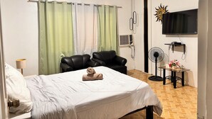 A hotel room with a bed, two chairs, a TV, a fan, and a wall-mounted air conditioner.