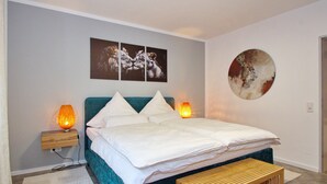 Ein Schlafzimmer mit einem turquoisefarbenen Bett, weißer Bettwäsche, zwei Nachttischlampen, einer Holzbank und Bildern an den Wänden.