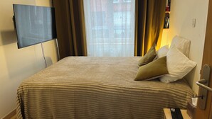 Ein Hotelzimmer mit Bett, Wandfernseher, Fenster mit Vorhängen und einer Tür mit Griff.
