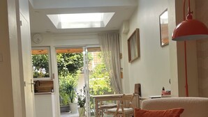 Ein großzügiger Wohnbereich mit einem Glastisch, einer Couch mit rotem Kissen und einer roten Stehlampe. Durch eine Glastür ist ein Gartenblick möglich.