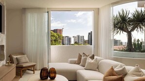 A modern living room with a stone fireplace, a comfortable sofa, and a large window offering a city view. - Cala by Khove (Burleigh Heads)