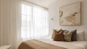 A bedroom with a bed, pillows, a window with sheer curtains, and a wall-mounted abstract painting. - Cala by Khove (Burleigh Heads)