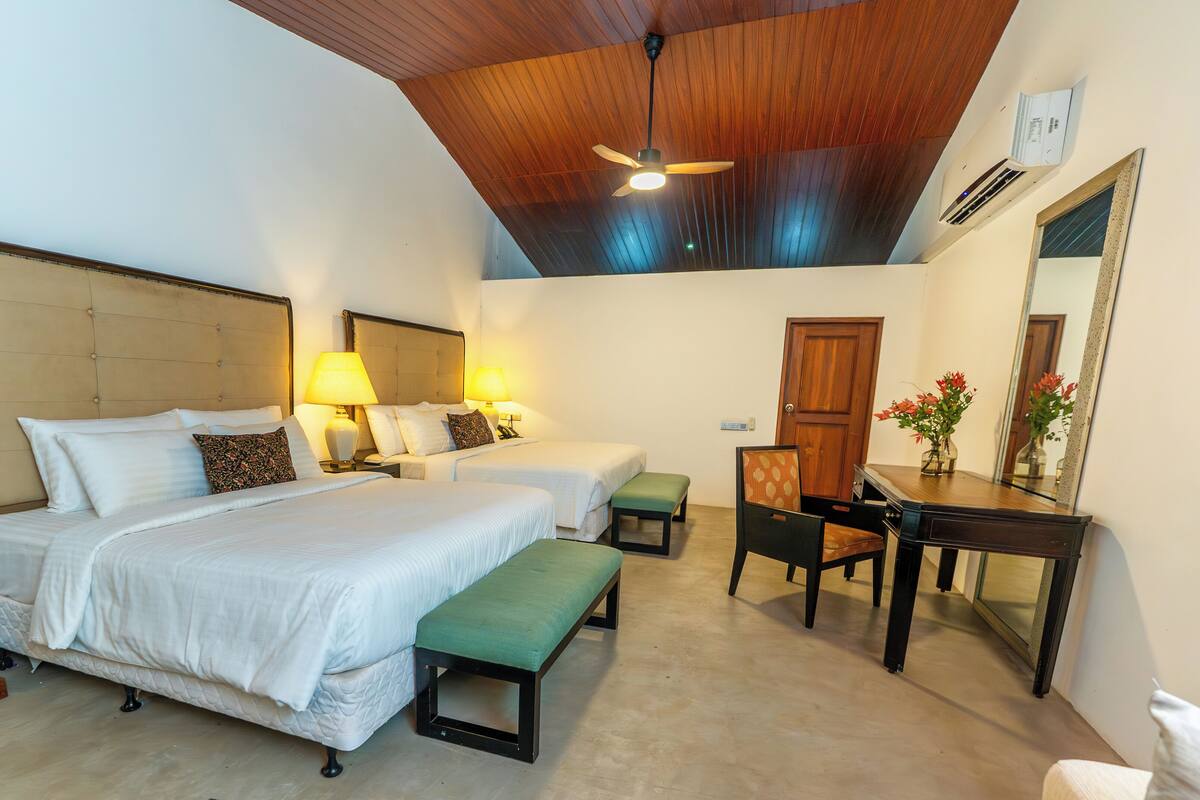 A hotel room with a large bed, two bedside tables with lamps, a green bench, a wooden desk with a mirror, and a ceiling fan.