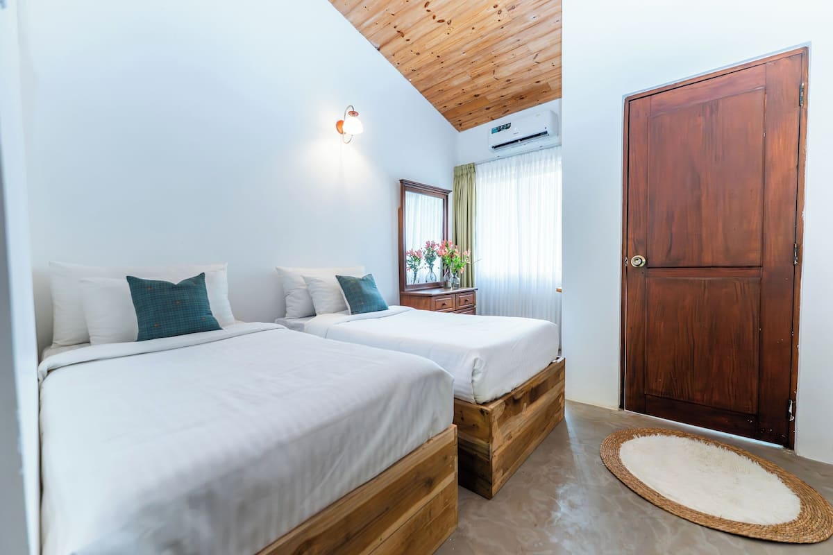 A bedroom with two beds, a wooden ceiling, an air conditioning unit, a wooden door, and a small rug.