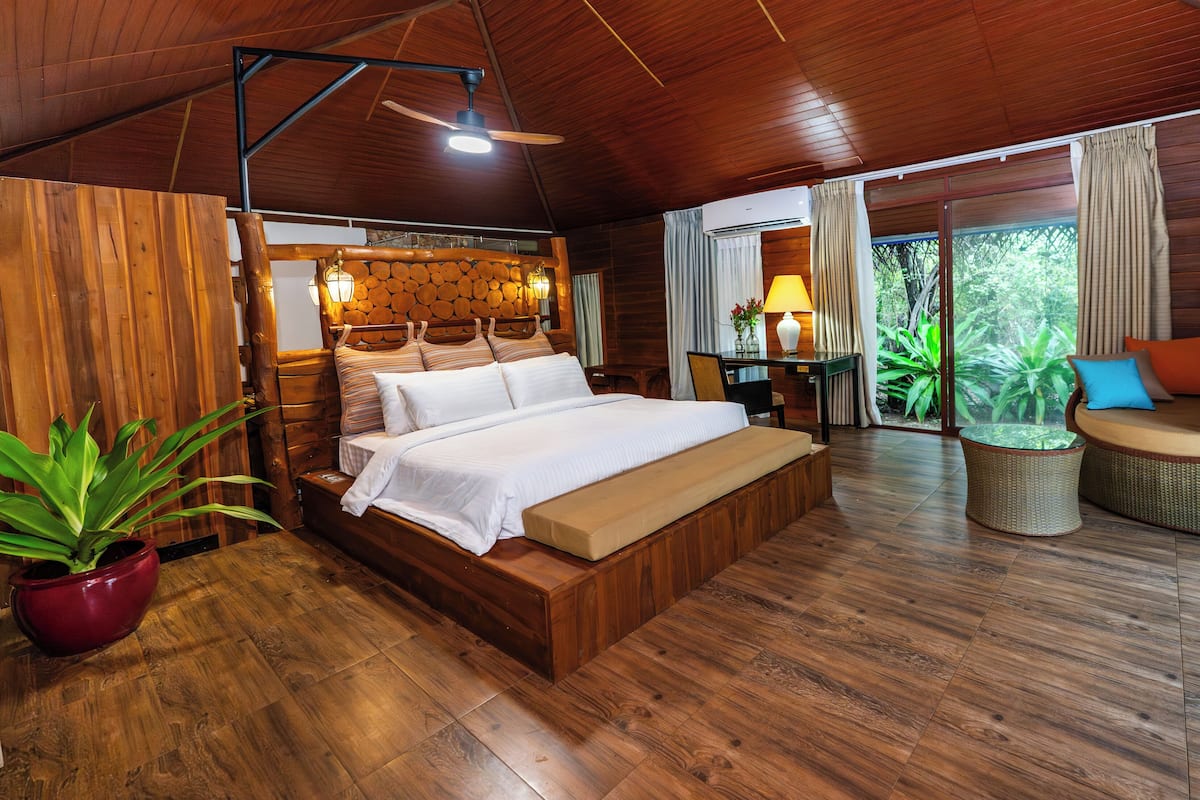 A wooden-themed bedroom with a bed, a seating area, and a view of greenery.