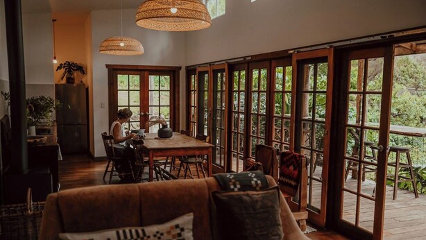 Un vaste espace de vie comprenant une grande table à manger en bois, un canapé en cuir marron agrémenté de coussins à motifs et de grandes fenêtres offrant une vue sur la verdure environnante.