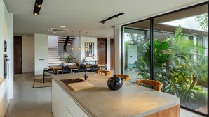A modern kitchen with a central island, wooden chairs, and a dining area with a view of indoor plants. - Villa Rosa by Pertama Stay (Kerobokan)