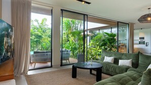 A modern living room with a green sofa, a round coffee table, and a large sliding glass door leading to a balcony with a view of lush greenery. - Villa Rosa by Pertama Stay (Kerobokan)