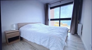 Een slaapkamer met een groot bed, een nachtkastje, een raam met gordijnen en uitzicht op bomen.