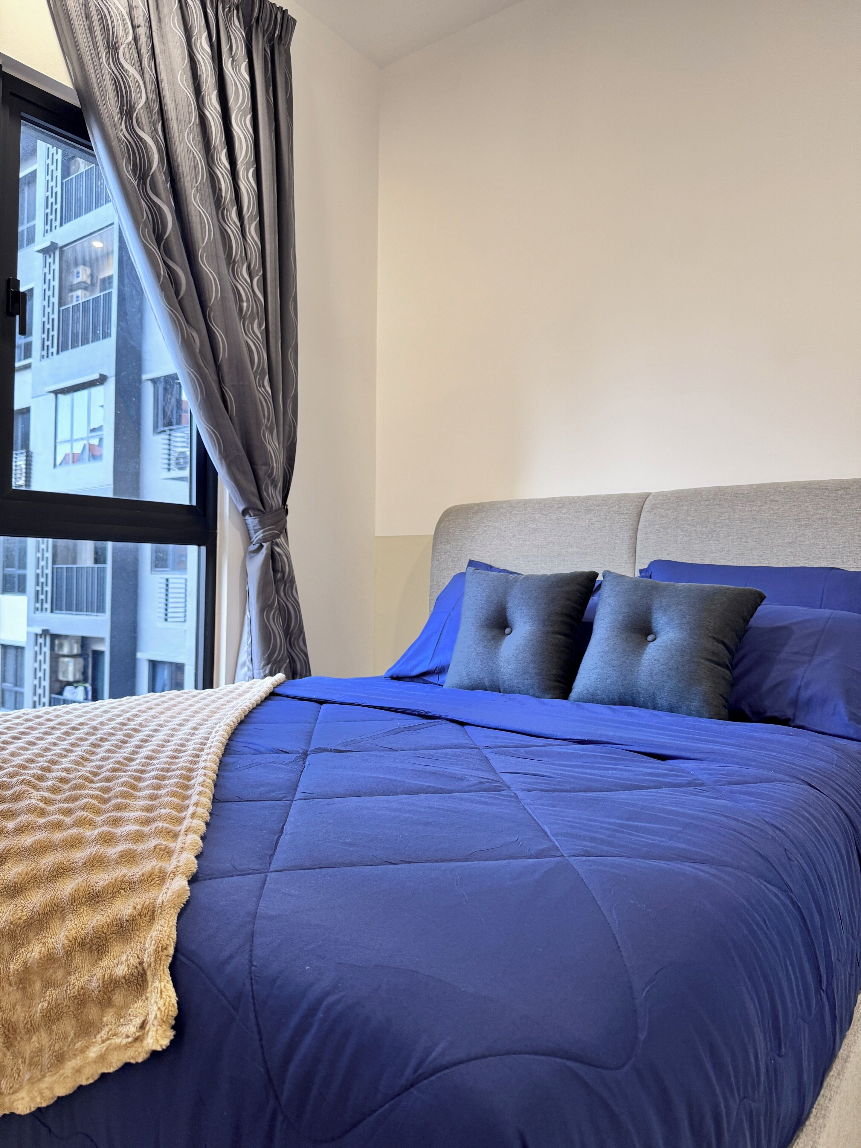 A neatly made bed with a blue duvet and grey pillows, a beige throw blanket, and a view of a building through a window with grey curtains.