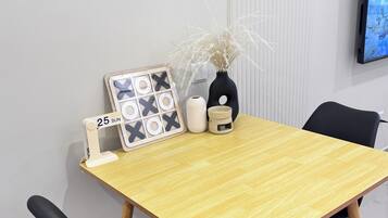 A dining table with a light wood finish, black chairs, a framed calendar, a vase with dried plants, and a wall-mounted TV.