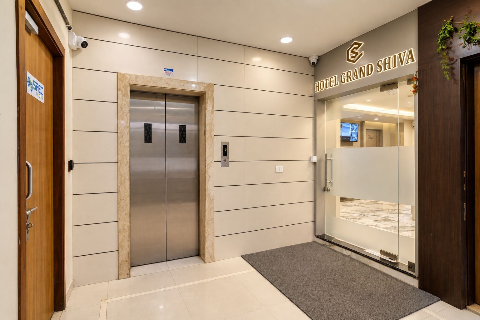 A hotel entrance with a modern elevator, a reception desk, and a sign for 'Hotel Grand Shiva.'