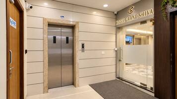 A hotel entrance with a modern elevator, a reception desk, and a sign for 'Hotel Grand Shiva.'