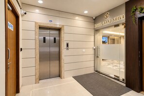 Een hotelingang met een moderne lift, een receptiebalie en een bord met de tekst 'Hotel Grand Shiva'.