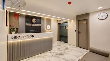 Uma área de recepção de hotel com um balcão de recepção identificado como 'RECEPTION', um relógio na parede e um letreiro 'HOTEL GRAND SHIVA'.