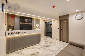 Un'area della reception di un hotel, con una scrivania reception che porta l'indicazione 'RECEPTION', una sveglia appesa al muro e un cartello che recita 'HOTEL GRAND SHIVA'.