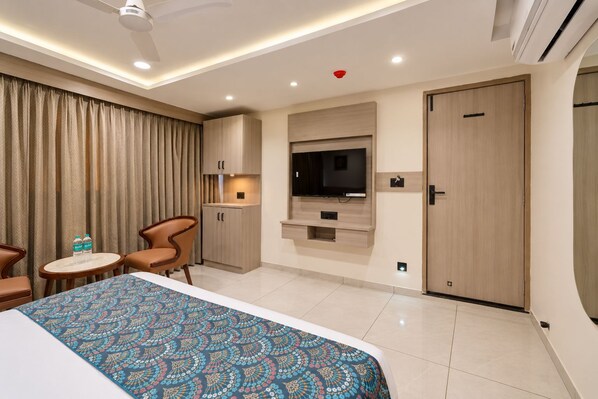 A hotel room with a bed, a TV mounted on the wall, a small table with chairs, and a wardrobe. - HOTEL GRAND SHIVA (Ahmedabad)
