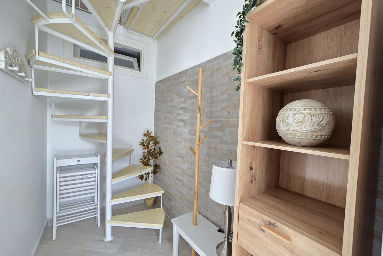 Et moderne interiør med en spiraltrappe, et træskab med en dekorativ skål og et hvidt bord med en lampe.