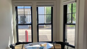 A room with a round table, two chairs, a pendant light, and large windows with a view of the street. - Great Island Inn - Standard Queen Guest Room (New Castle)