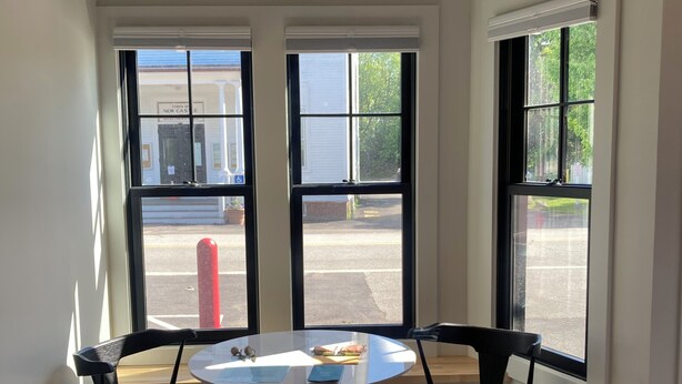 Una habitación con una mesa redonda, dos sillas, una lámpara colgante y ventanas amplias con vista a la calle.