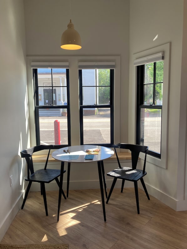 Una habitación con una mesa redonda, dos sillas, una lámpara colgante y ventanas amplias con vista a la calle.