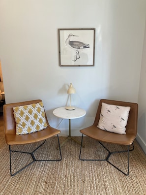 Una habitación con dos sillas marrones, una mesita redonda con una lámpara y un cuadro de aves en la pared.