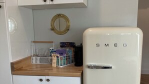Una cocina con armarios blancos, encimera de madera, un refrigerador blanco con la etiqueta 'SMEG', un espejo redondo pequeño y una cafetera.
