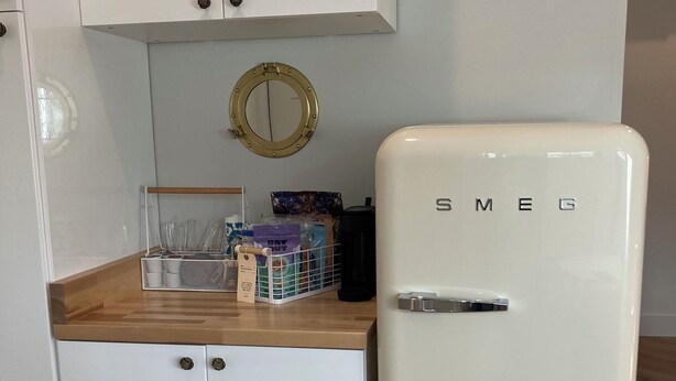 A kitchen with white cabinets, a wooden countertop, a white refrigerator labeled 'SMEG', a small round mirror, and a coffee maker. - Great Island Inn - Standard Queen Guest Room (New Castle)