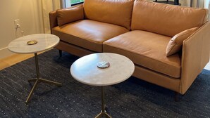 Un salon moderne avec un canapé en cuir marron, deux petites tables rondes, une moquette foncée et un coin repas à l’arrière-plan.