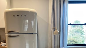 Une cuisine compacte avec un réfrigérateur Smeg blanc, une chaise en bois avec un coussin blanc et une fenêtre avec des rideaux.