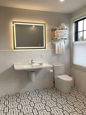 Et moderne badeværelse med hvid vask, toilet og spejl med indbygget lys. Der er et håndklædetørrestativ med håndklæder og toiletpapirholder. Gulvet har et sort-hvidt geometrisk flisemønster.