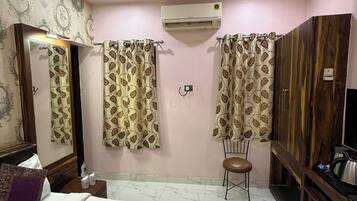 A hotel room with a bed, a ceiling fan, an air conditioning unit, a wardrobe, a chair, and a small table with a kettle.