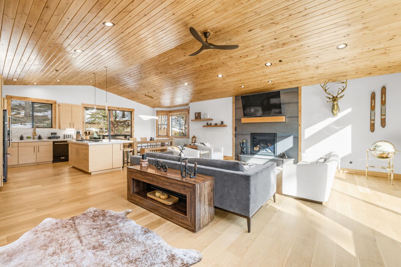 Un vaste espace de vie comprenant un grand canapé gris, une table basse en bois et une cheminée. La cuisine est dotée de meubles en bois et d’une grande fenêtre donnant sur un paysage enneigé.