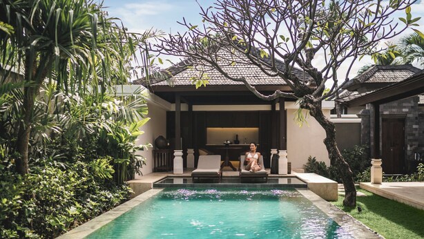 A poolside area with a person sitting on a lounge chair, surrounded by lush greenery and a traditional building with a tiled roof. - Taman Estate by Ekosistem (Seminyak)
