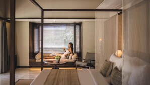 A modern hotel room with a large bed, a sitting area with a sofa and armchairs, a coffee table, and a large window with blinds. - Taman Estate by Ekosistem (Seminyak)