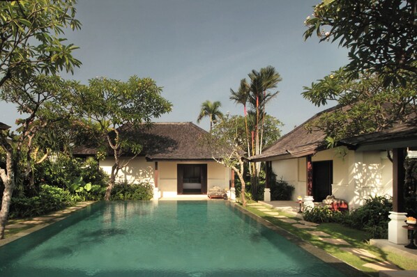 A pool area with a house in the background, surrounded by lush greenery and palm trees.