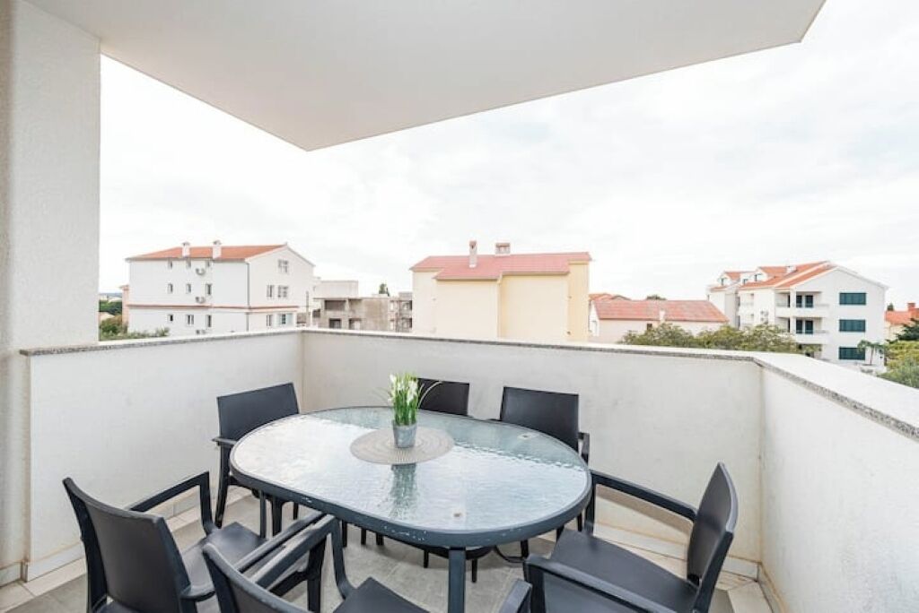 Un balcón con una mesa redonda de vidrio y seis sillas, con vista a una zona residencial con edificios.