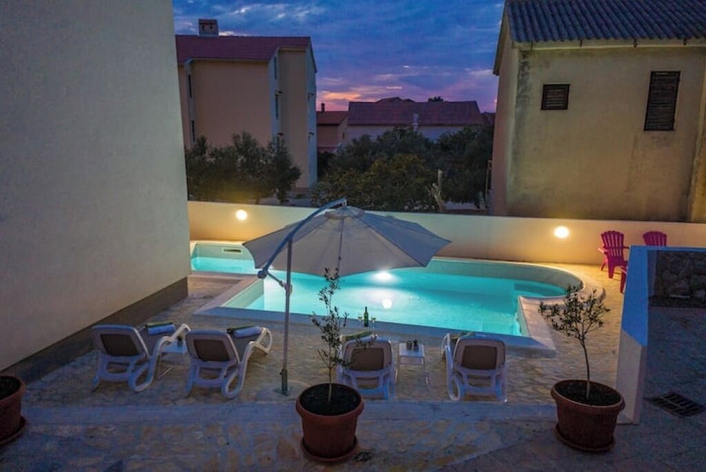 Una piscina exterior con sillones de descanso y sombrillas, rodeada de edificios y plantas en macetas.