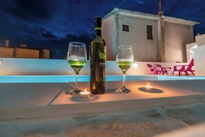 Una terraza con una botella de vino y dos copas sobre una mesa, una vela pequeña y un edificio al fondo.