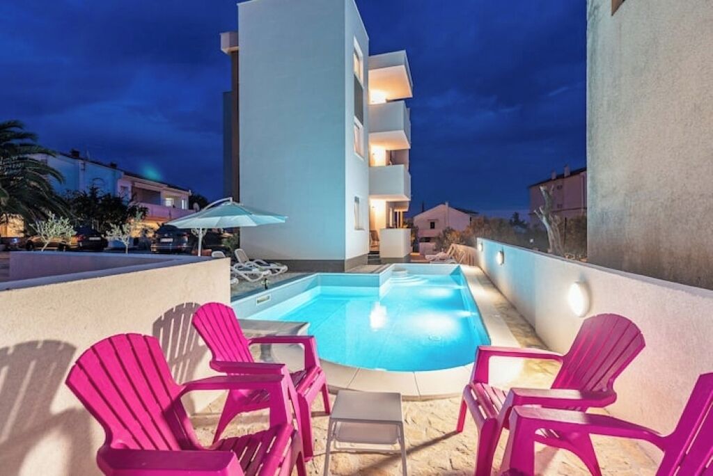Zona de piscina al terrat amb piscina, gandules roses i un edifici blanc al fons.