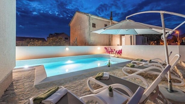 Ein Swimmingpool mit Liegestühlen und einer überdachten Sitzgelegenheit im Dämmerlicht.