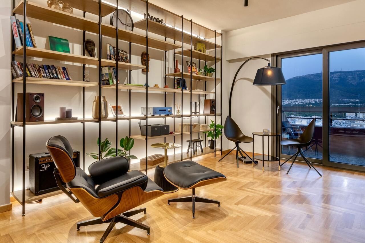 A modern living room with a wooden floor, a black leather sofa, a black ottoman, a bookshelf with various items, a small table with a lamp, and a view of a cityscape through large windows.