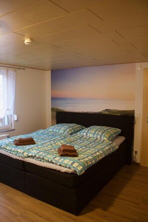 Une chambre à coucher avec un lit, une fenêtre avec des rideaux, une fresque murale représentant une plage et une porte.