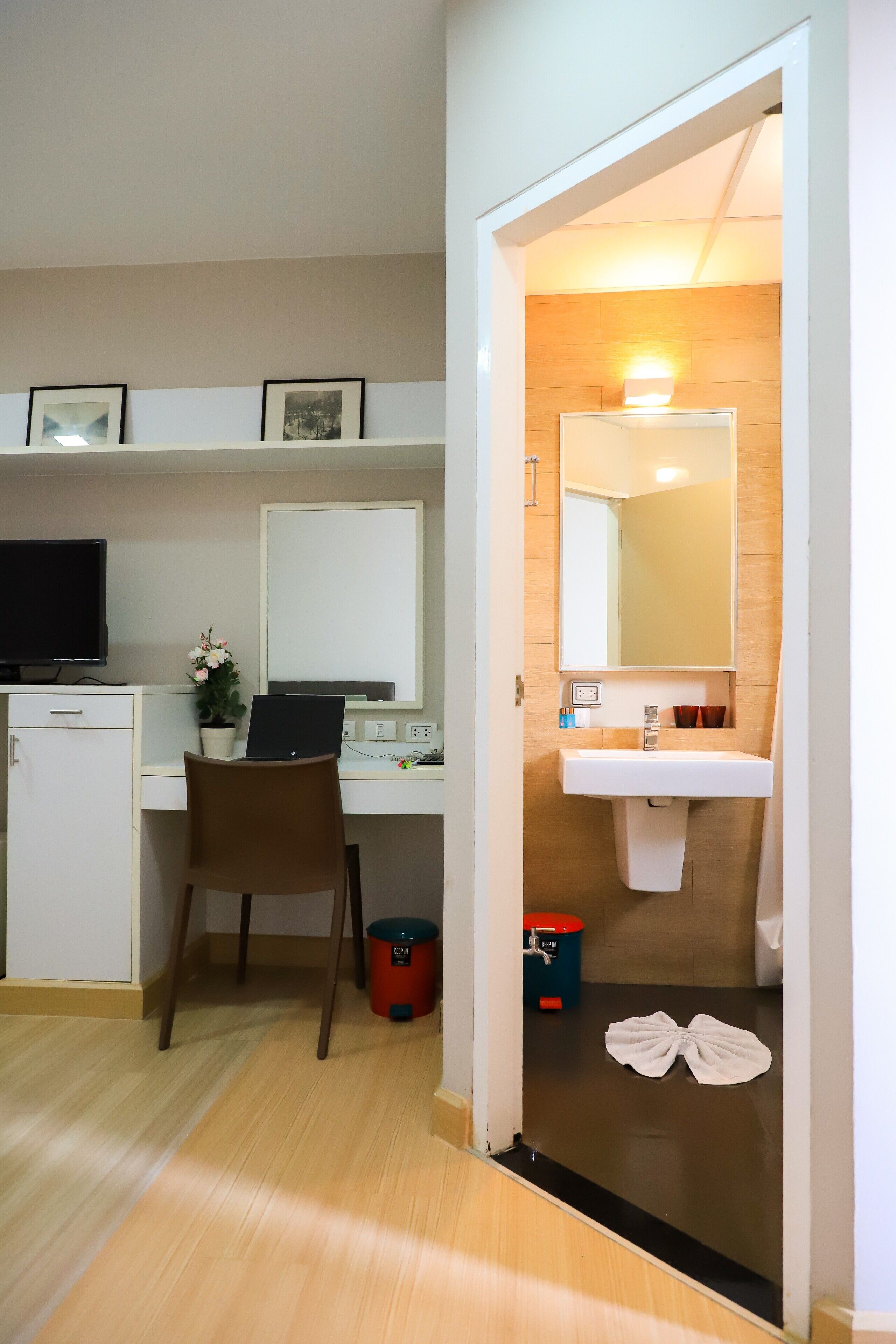 A hotel room with a desk, chair, computer, and a small cabinet. There is a bathroom with a sink and mirror.