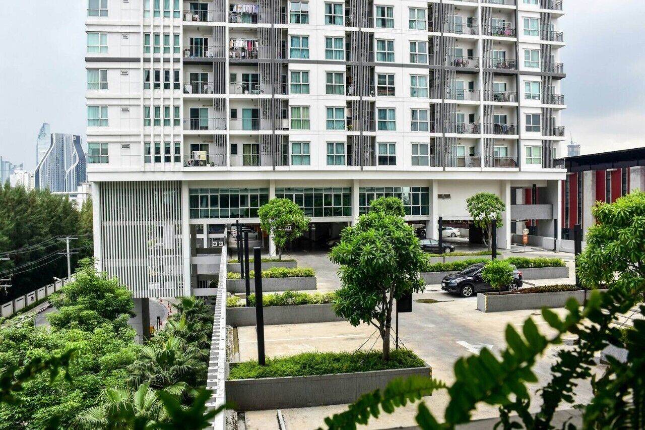 A modern multi-story building with balconies, a parking area, and surrounding greenery.